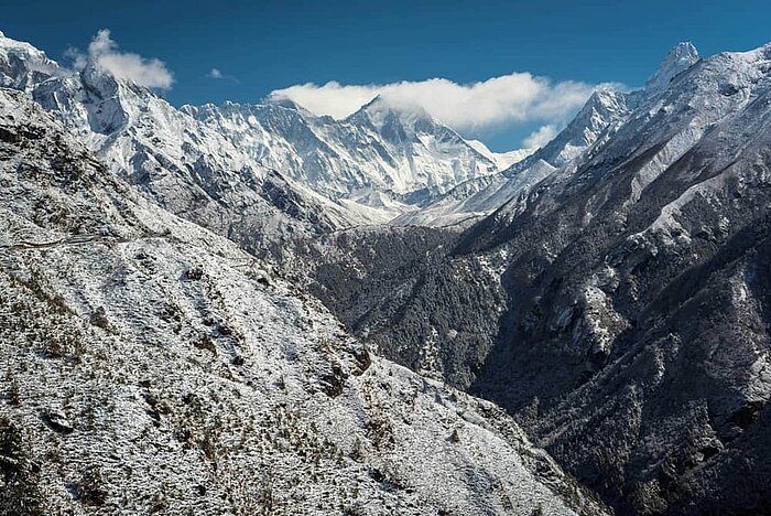 Ein atemberaubendes Gebirgspanorama zeigt schneebedeckte Gipfel, die hoch in den klaren blauen Himmel ragen. Zwischen den Bergen erstreckt sich ein grünes Tal. Die Sonne scheint auf die Landschaft und hebt die texturierten Felsen und die verschneiten Hänge hervor.